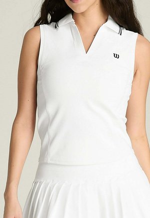 Donna che indossa una polo bianca senza maniche con colletto nero bordato e gonna bianca plissettata.