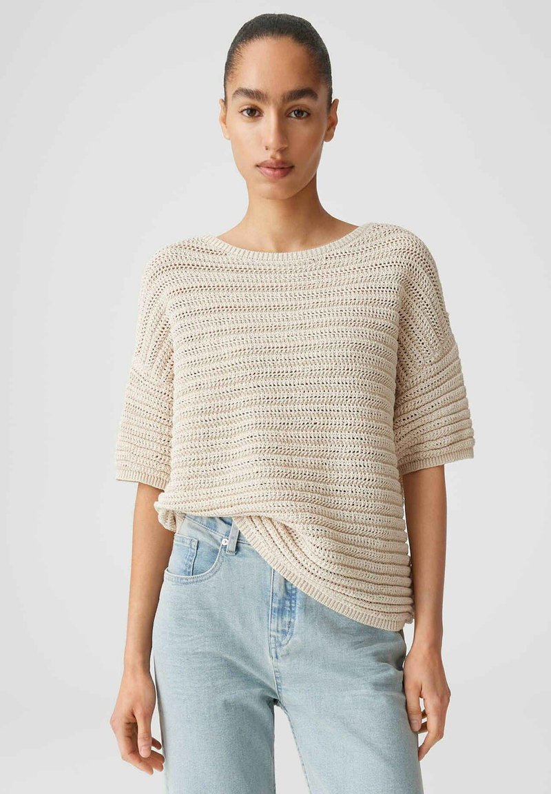 Junge Frau mit zurückgebundenem Haar, die einen beigen Kurzarm-Strickpullover und hellblaue, hoch taillierte Jeans vor einem schlichten Hintergrund trägt.