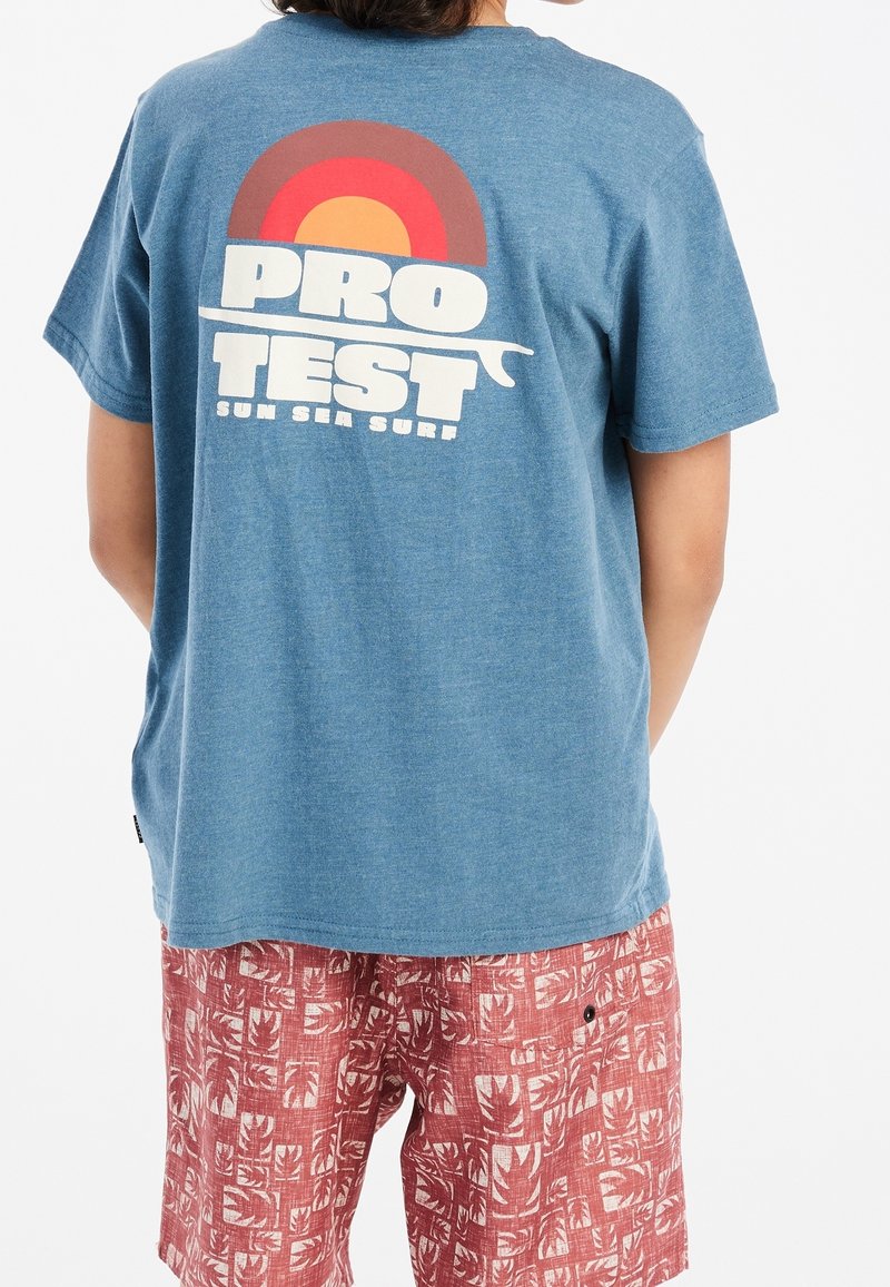 Personne portant un t-shirt bleu avec "PRO TEST SUN SEA SURF" et un graphisme de soleil rouge-orange, associé à un short rouge à motifs.