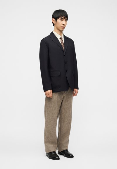 Άνδρας που φοράει μαύρο σακάκι, μπεζ γραβάτα με σχέδια, κρεμ πουκάμισο, φαρδιά μπεζ παντελόνια και μαύρα παπούτσια, στέκεται μπροστά σε απλό φόντο.