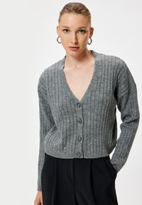 Cárdigan gris de canalé con un escote en V profundo, que presenta cinco rayas verticales, cierre de botones y mangas largas. Textura suave y diseño ajustado.