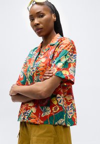 Γυναίκα με σταυρωμένα τα χέρια φορόντας πολύχρωμο κόκκινο πουκάμισο με λουλούδια και μουσταρδί παντελόνι, με γυαλιά να ακουμπούν στο κεφάλι της, μπροστά σε απλό φόντο.