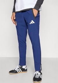 Μπλε αθλητικό παντελόνι με φερμουάρ στις τσέπες, λογότυπο Adidas και ταμπελάκι στο μηρό. Κατασκευασμένο από απαλό, ελαφρύ υλικό.