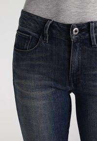 Vaqueros de denim azul oscuro con corte slim, que cuentan con un bolsillo frontal, cierre de botón y detalles de costura visibles en la cinturilla.