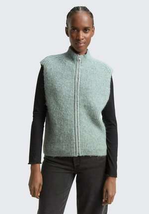 Hellgrüner gestrickter Weste mit hohem Kragen, durchgehendem Reißverschluss und ohne Ärmel, getragen über einem schwarzen Langarmoberteil. Strukturiertes Oberflächendesign.