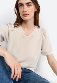 Blouse beige à manches courtes avec un décolleté rond et des manches bouffantes, réalisée en tissu lisse. Le mannequin est assis, portant un jean en denim.