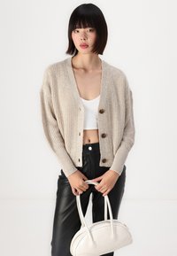 Beige pletený cardigan s V-výstřihem, třemi velkými hnědými knoflíky a žebrovanými manžetami, kombinovaný s černými koženými kalhotami a bílou kabelkou.