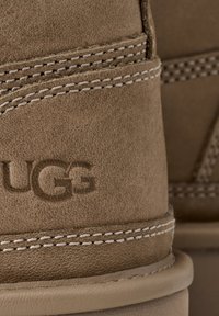 Gros plan sur le talon d'une botte UGG en daim marron montrant des coutures détaillées et un logo de marque en relief.