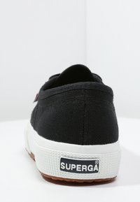 Zapatillas de lona negras con suela de goma blanca, acabado texturizado, logo en relieve en el talón y un interior marrón en contraste.