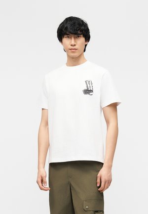 Jeune homme aux cheveux noirs porte un t-shirt blanc à manches courtes avec un petit motif noir « Motel Service » et un pantalon cargo vert olive.