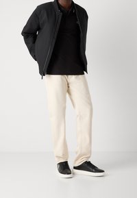 Černá bomber bunda přes černé polo tričko, doplněná krémovými kalhotami a černými teniskami s bílými podrážkami. Jednoduchý design, jednofarebné.