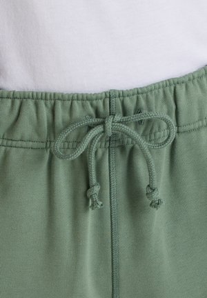 Vita elasticizzata con coulisse annodata su pantaloni da jogging verde chiaro su sfondo bianco.