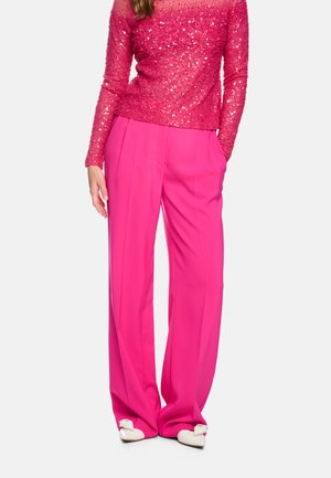 Woman wearing bright pink wide-leg pants, a long-sleeve sparkly pink top, and white shoes with bows, standing against a plain background.