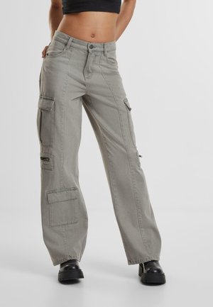 Femme portant un pantalon cargo gris clair avec plusieurs poches et des bottes à plateforme noires, debout devant un fond clair uni.