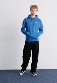 Hoodie azul com um bolso frontal e cordões, combinado com calças largas pretas e sapatilhas cinzentas com detalhes pretos. Tecido texturizado.