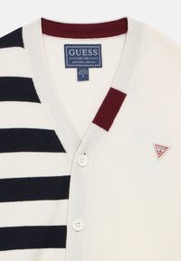 Κρεμ ζακέτα με οριζόντιες ναυτικές και λευκές ρίγες, V-λαιμόκοψη, τρία κουμπιά και μια ετικέτα με λογότυπο με κόκκινη λεπτομέρεια στην αριστερή πλευρά.