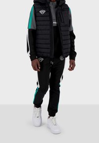 Svart pufferväst med huva över grå sweatshirt och svarta träningsbyxor. Byxorna har turkosa och vita ränder. Vita sneakers.