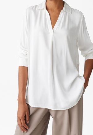 Kvinde iført en løs hvid bluse med lange ærmer og krave samt beige bredbenede bukser, med den ene hånd i lommen, på hvid baggrund.