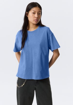 Jeune personne aux longs cheveux noirs, portant une chemise bleue à manches courtes et amples ainsi qu'un pantalon noir avec un accessoire en chaîne argentée.