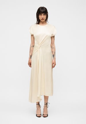 Kvinde med mørkt hår og tatoveringer iført en off-white asymmetrisk midikjole med knudedetalje, kombineret med sorte snørede høje hæle.