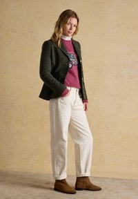 Vrouw draagt een donkergroene blazer, roze trui met autodesign, witte broek en bruine enkellaarzen, staand tegen een beige achtergrond.