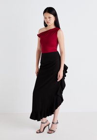 Body rosso a spalla unica abbinato a una maxi gonna nera con accenti arricciati. Sandali neri con tacco alto completano il look. Texture lisce.