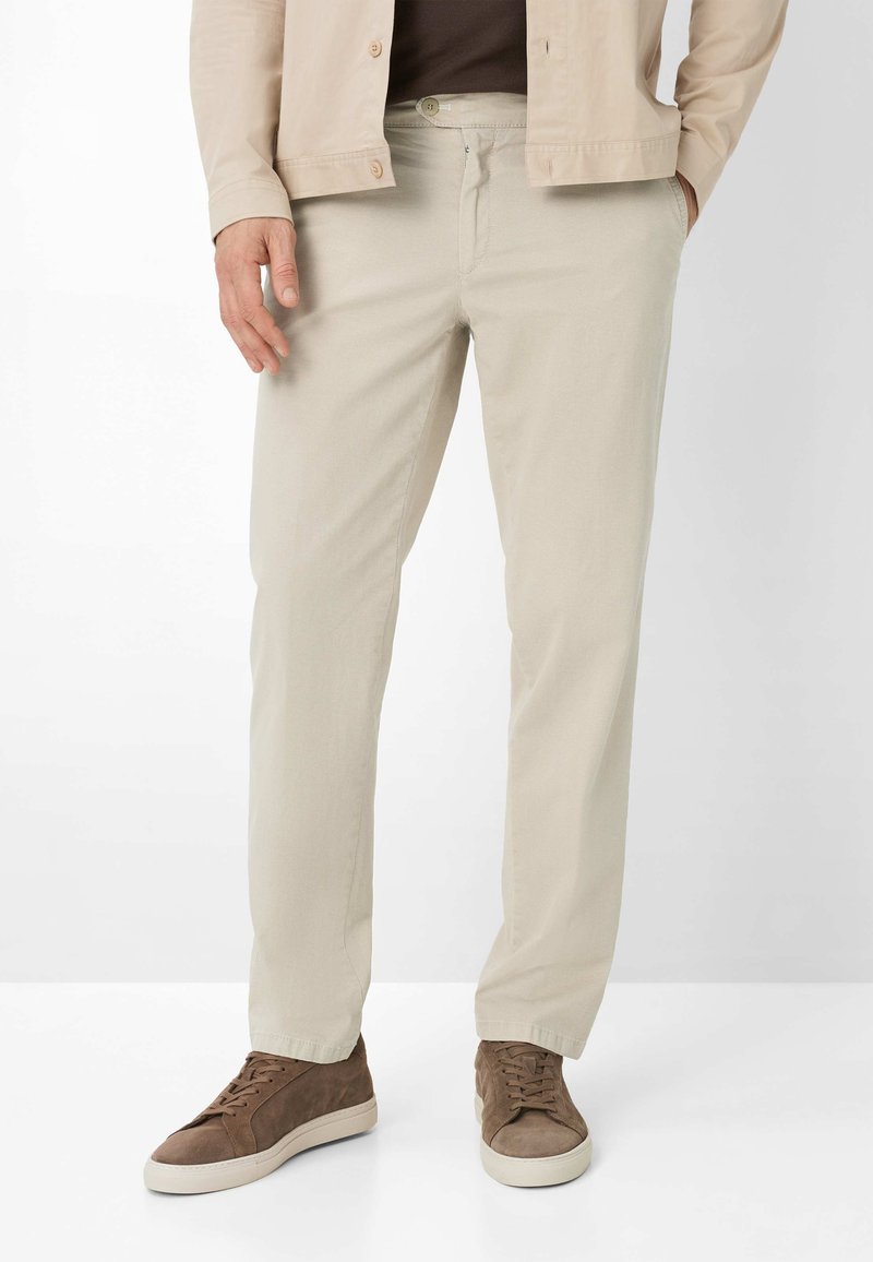 Mann trägt beige Hose, braune Wildledersneaker und eine hellbeige Jacke, die Hände in den Taschen, vor weißem Hintergrund.