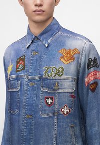 Homme portant une veste en denim bleue avec plusieurs patchs brodés colorés et poches poitrine, visible des épaules jusqu'au milieu du torse.