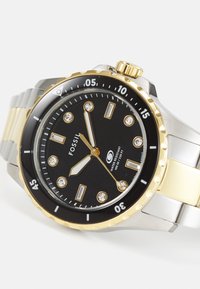 Fossil ur med sort urskive, bånd i guld og sølv metal, lysende timeindikatorer og en roterende bezel. Vandafvisende op til 100 meter.