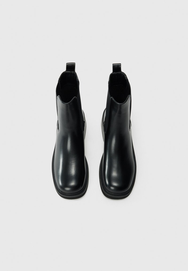 Bottines en cuir noir avec une finition lisse, des panneaux latéraux élastiques et des tirettes au talon, présentant une pointure arrondie et une semelle plate.