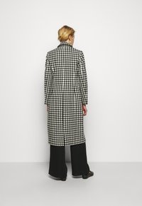 Lange jas met een zwart-wit ruitpatroon, voorzien van een reverskraag en een split aan de achterkant. De stof lijkt textuur en structuur te hebben.