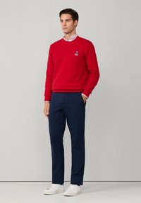 Sudadera roja con cuello redondo y logo bordado en azul, combinada con pantalones de marina y zapatillas blancas; tejido suave, ajuste casual.