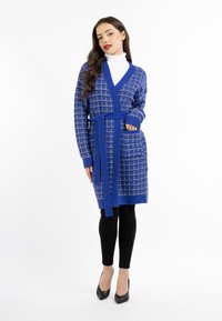 Kék kötött kardigán kockás mintával, szűkített övvel, két zsebbel és bordázott mandzsettával, fekete leggings és magassarkú cipő fölött viselve.