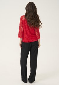 Haut en dentelle rouge à manches trois-quarts, avec un ourlet festonné, associé à un pantalon noir à jambes larges. La dentelle présente un motif floral.