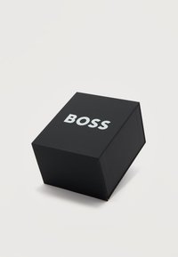 Μαύρο κουτί σε σχήμα κύβου με τη λέξη "BOSS" σε λευκά κεφαλαία γράμματα στην κορυφή, τοποθετημένο πάνω σε ανοιχτογκρι επιφάνεια.