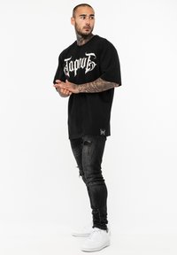 Črn oversize majica s belim grafičnim napisom, v kombinaciji s strganimi črnimi jeans hlačami in belimi supergami. Model pozira bočno.