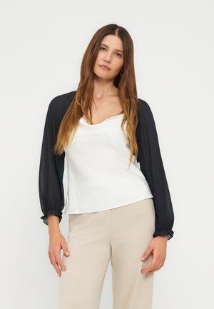 Femme aux longs cheveux bruns portant un haut blanc avec des manches longues transparentes noires et un pantalon beige, debout devant un fond gris uni.