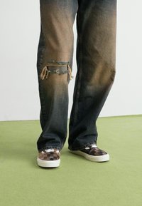 Mørke denimjeans med en falmet, slitt finish og et stort hull ved kneet, parret med brune sko med sjakkmønster.