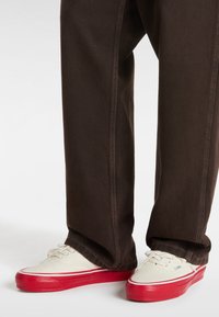 Die Canvas-Sneaker haben ein weißes Obermaterial mit roten Gummisohlen, kombiniert mit dunkelbraunen Hosen, die kontrastierende Nähte aufweisen.