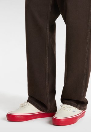 Canvas sneakers feature a white upper with red rubber soles, paired with dark brown pants showcasing contrasting stitching details.