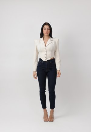Mujer de pie con una blazer blanca ajustada con hombros abullonados, jeans oscuros ajustados y sandalias de tacón alto transparentes sobre un fondo liso.