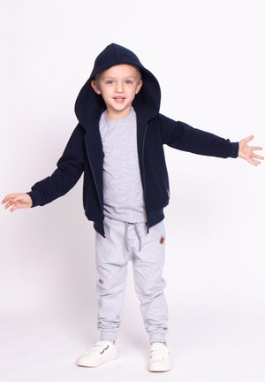 Uśmiechnięty młody chłopiec w granatowej kurtce z kapturem, szarej koszulce i spodniach, białych trampkach, stojący z rozłożonymi ramionami na jednolitym tle.
