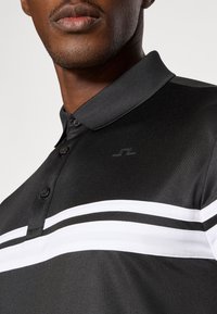 Μαύρο πουκάμισο πόλο με διακριτική γυαλάδα, διαθέτοντας μαύρο γιακά, κουμπότρυπα και λευκές ρίγες διαγωνίως στο στήθος.