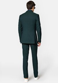 Zielony garnitur o dopasowanym kroju, teksturowany materiał, z pojedynczym rozcięciem z tyłu oraz białymi trampkami. Niebieska koszula w białe kropki widoczna przy kołnierzyku.