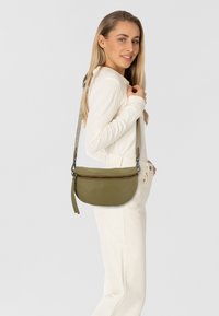 Borsa a tracolla in pelle verde oliva con forma arrotondata, chiusura con zip e tracolla testurizzata con logo del marchio. Il modello indossa un top e pantaloni di colore crema.
