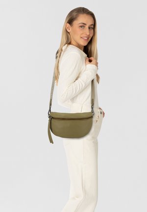 Borsa a tracolla in pelle verde oliva con forma arrotondata, chiusura con zip e tracolla testurizzata con logo del marchio. Il modello indossa un top e pantaloni di colore crema.
