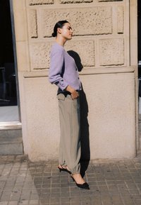 Frau mit zurückgebundenem Haar trägt einen lavendelfarbenen Pullover, beige lockere Hosen und schwarze High Heels, steht mit geschlossenen Augen an einer strukturierten Wand.