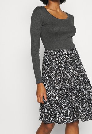 Femme portant un haut ajusté à manches longues gris foncé et une jupe noire à imprimé floral blanc sur un fond clair uni.