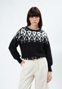 Schwarzer Strickpullover mit weißem geometrischem Muster am Schulterbereich, Rundhals-Ausschnitt und gerippten Bündchen, getragen mit cremefarbenen Hosen und einem Gürtel.