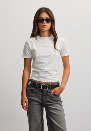 Kvinde iført sorte solbriller, hvid tætsiddende t-shirt, sort bælte og falmede sorte jeans, stående med den ene hånd i lommen mod en ensfarvet baggrund.